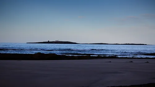 This landscape photograph captures the Farne Islands viewed from a wide stretch of beach in Northumberland, England, United Kingdom. Taken in the afternoon during the autumn season, the image showcases the calm water of the North Sea with gentle waves rolling towards the sandy shoreline. The distant Farne Islands are clearly visible across the water, and atop the islands, lighthouses stand as prominent landmarks, highlighting their maritime significance. The photograph effectively blends natural elements like the beach and sea with the historic lighthouses of the Farne Islands, presenting a scenic coastal setting typical of Northumberland, England in the United Kingdom.